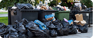 poubelle De grandes piles de sacs poubelles et de déchets en vrac débordent des bennes à ordures sur le sol, tandis que des cartons et divers déchets sont éparpillés dans un espace extérieur.